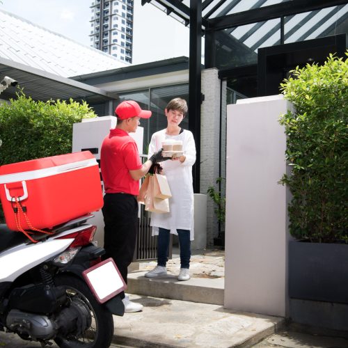 Asian delivery man in red uniform delivering  groceries bag of food, fruit, vegetable and drink to woman recipient at home