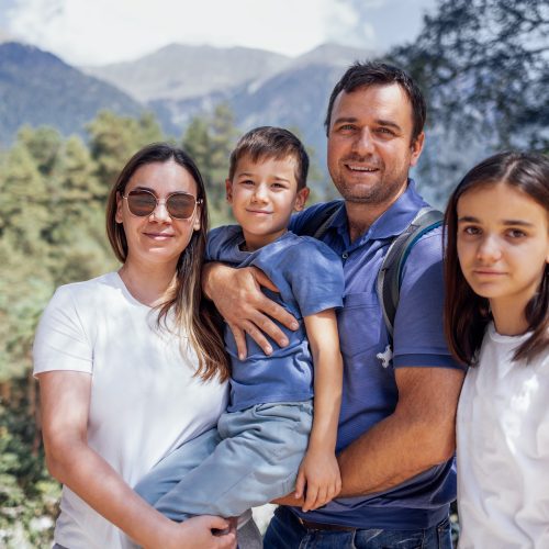 Happy family on background of mountain landscape. Young married couple with their son and daughter are walking in forest. Smiling tourists enjoy nature and travel. Mom, dad and their kids are in park.