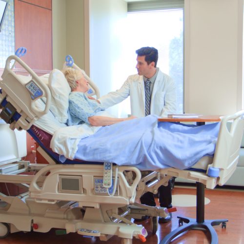 Male Doctor Wearing White Coat Visiting Senior Female Patient In Hospital Bed Giving Her Good News