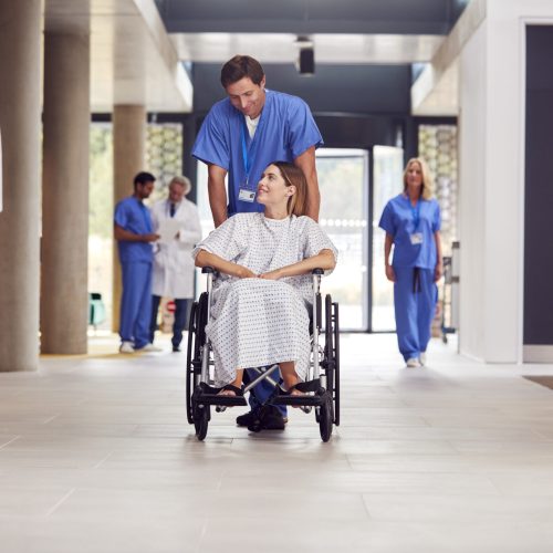 Male Nurse Wearing Scrubs Pushing Female Patient In Wheelchair Through Hospital Building