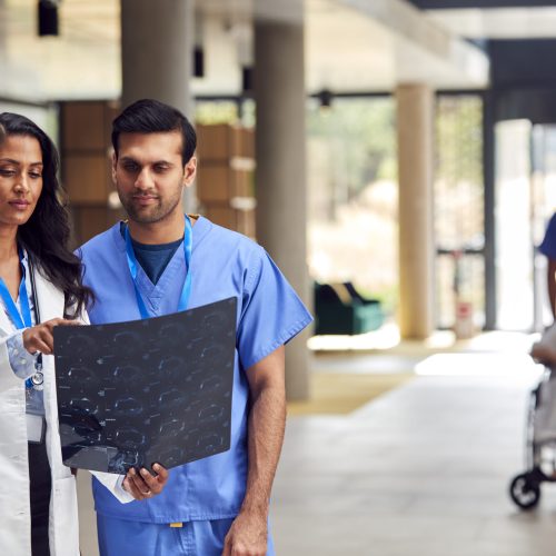 Two Medical Staff In White Coats And Scrubs With Patient Scans Having Informal Meeting In Hospital